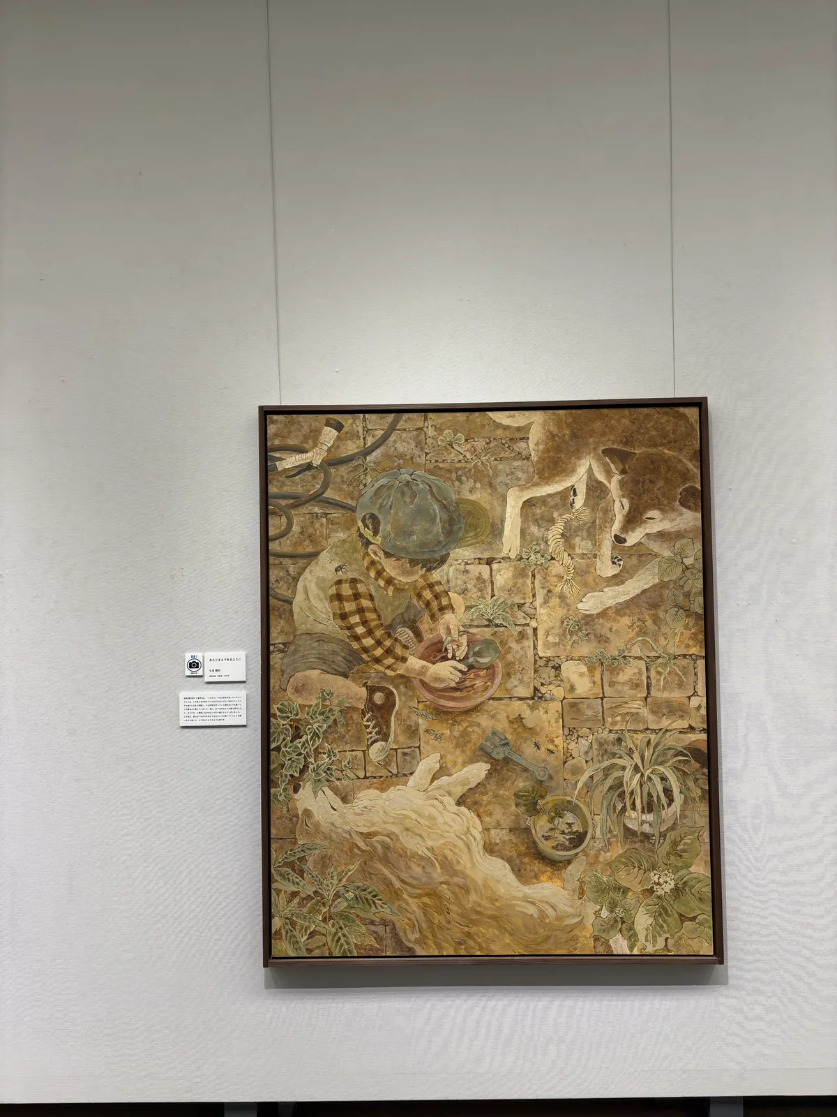 展示室的一面牆，掛著一幅以褐色為主調的大幅畫作，畫作右側是一張小型的作品標籤，牆面是帶有細緻紋理的白色壁布，下方是深色的踢腳與灰色地毯