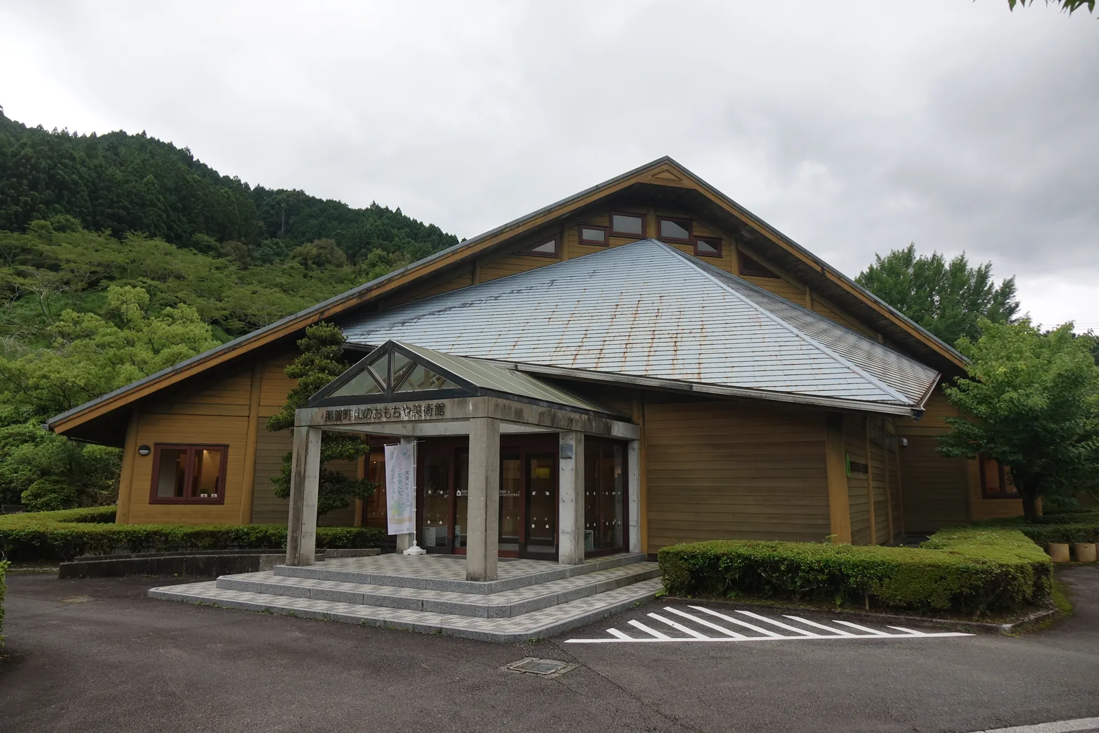 那賀町山之玩具美術館的外觀，一座覆有金屬板大屋頂的木造外觀建築，正面是一座混凝土柱支承的山牆形門廊，門廊上方是館名，左右兩翼是木板外牆，前方是車道與停車格