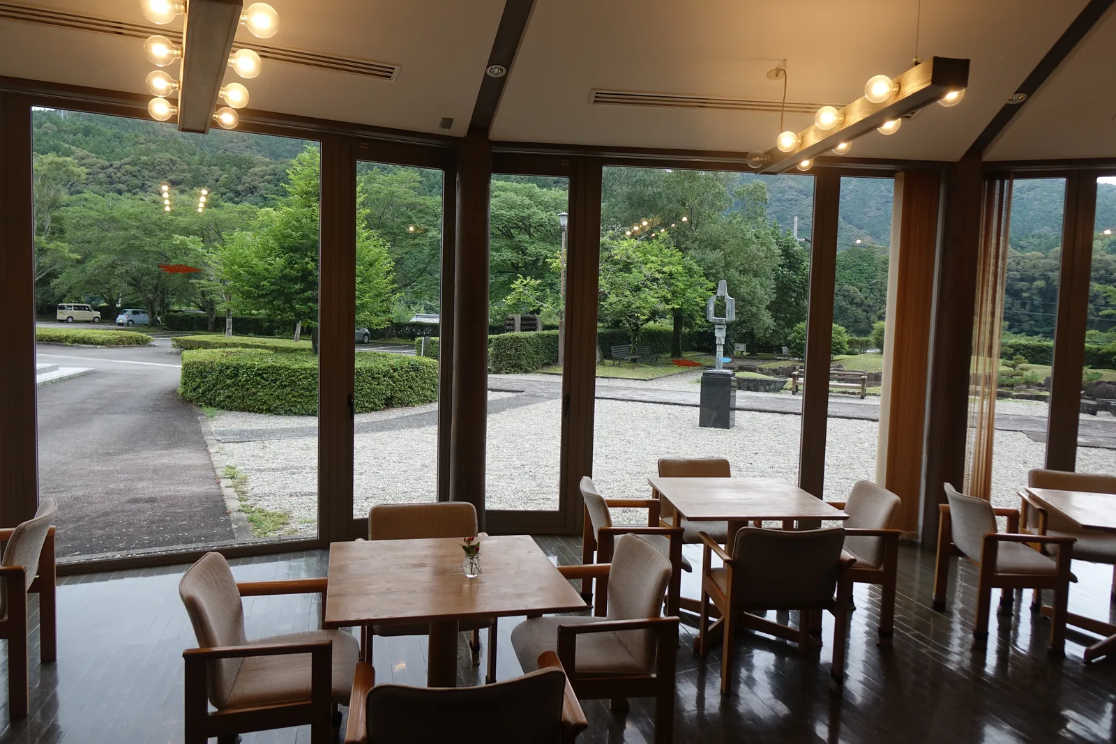 lounge 的內部，木框的整面玻璃圍成多角形的外牆，室內排列著木桌與扶手椅，天花垂下一排球形燈泡的吊燈，玻璃外是砂礫廣場、修剪整齊的綠籬與一座雕塑，遠處是山