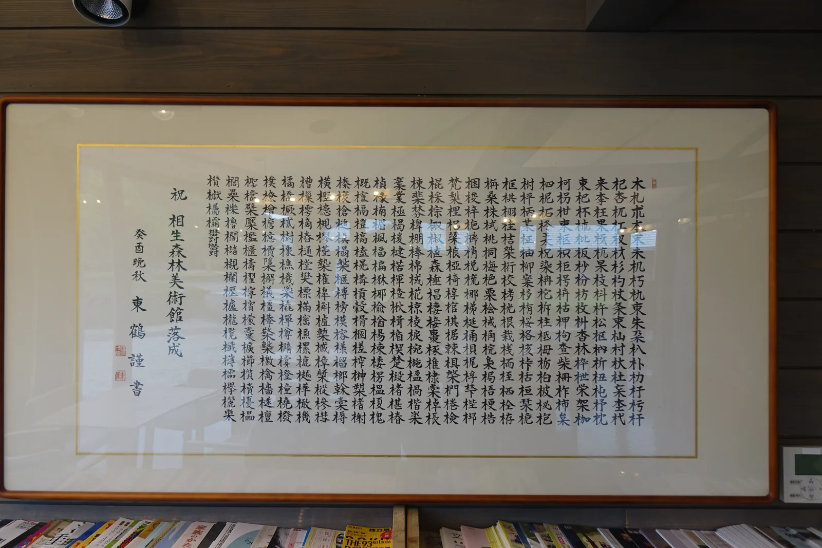 館內牆上的一幅大型書法額，白底上以楷書密密寫滿以木字旁構成的漢字，左側落款為祝相生森林美術館落成與癸酉晚秋，額下是一排擺放觀光刊物的書架