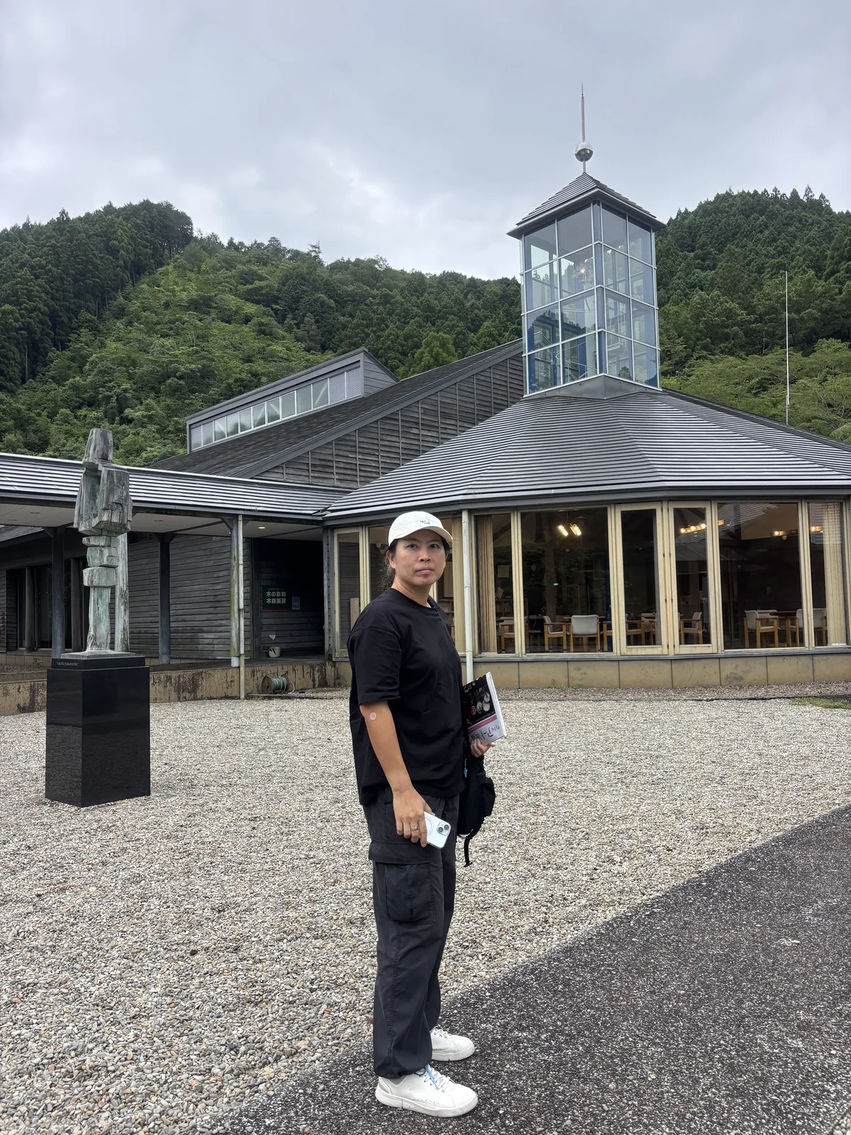 美術館前的砂礫廣場，前景一名參觀者站立，後方是美術館的灰色大屋頂、玻璃塔屋與多角形的 lounge，左側台座上立著一座雕塑，背後是杉林覆蓋的山