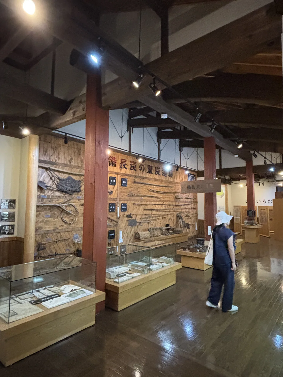 紀州備長炭發見館展示室，一排紅色方柱撐起深色木梁，梁下以細鋼棒斜拉構成屋架，左側木板牆上掛滿燒炭工具，下方是玻璃展示櫃，一位參觀者站在右側