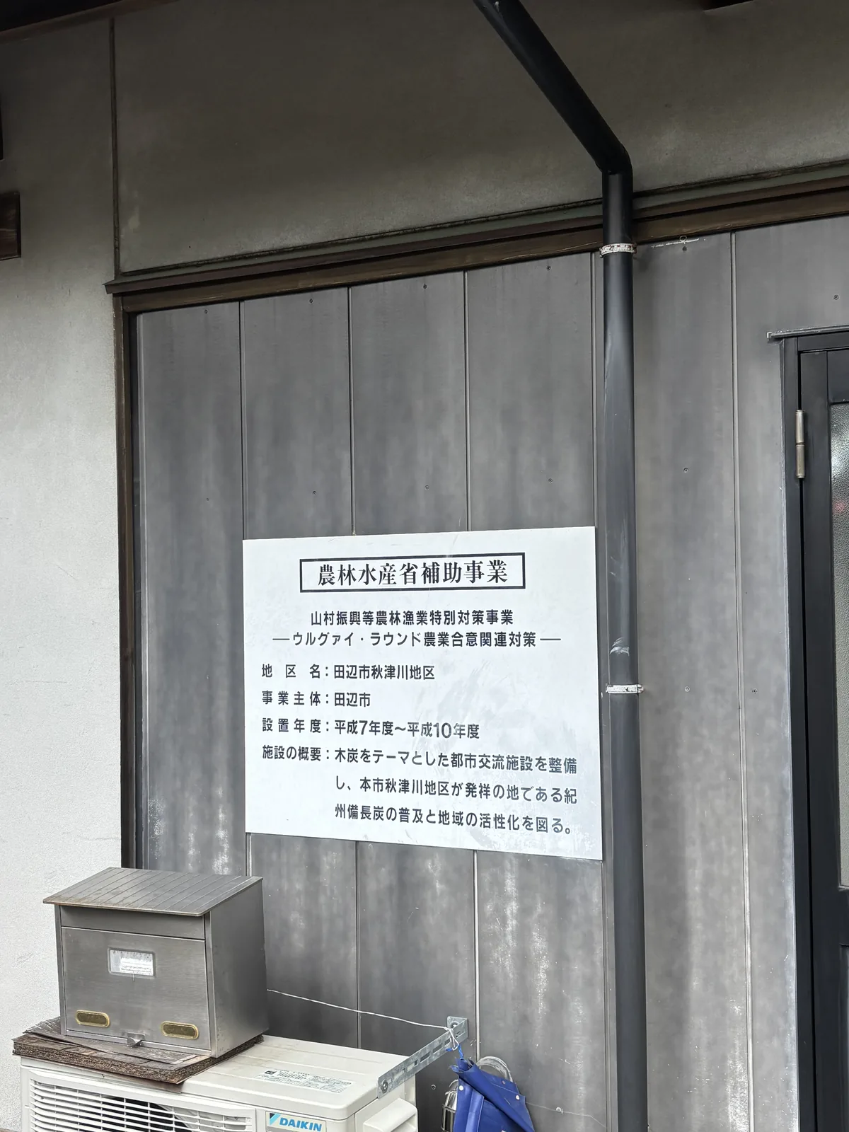 館舍外牆上的補助事業標示板，白底黑字寫著農林水產省補助事業、事業名稱、地區名、事業主體、設置年度與施設概要，背後是深灰色的縱向板材外牆與一根落水管，下方是冷氣室外機