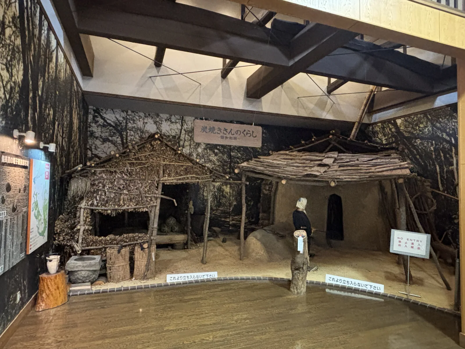 展示室一角重現的炭窯與燒炭小屋，左側是茅草屋頂的小屋，右側是樹皮屋頂的窯場與一尊人偶，上方是深色的木梁與白色天花，背景是描繪山林的壁畫