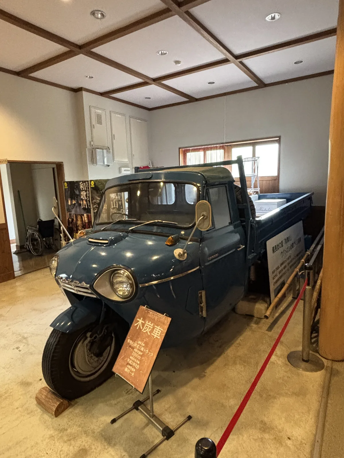 一個天花為白色格子天花的房間，中央展示一輛藍色的三輪木炭車，前方立著寫有木炭車的說明牌，以紅繩圍起，後方是木框的格子窗與幾扇門
