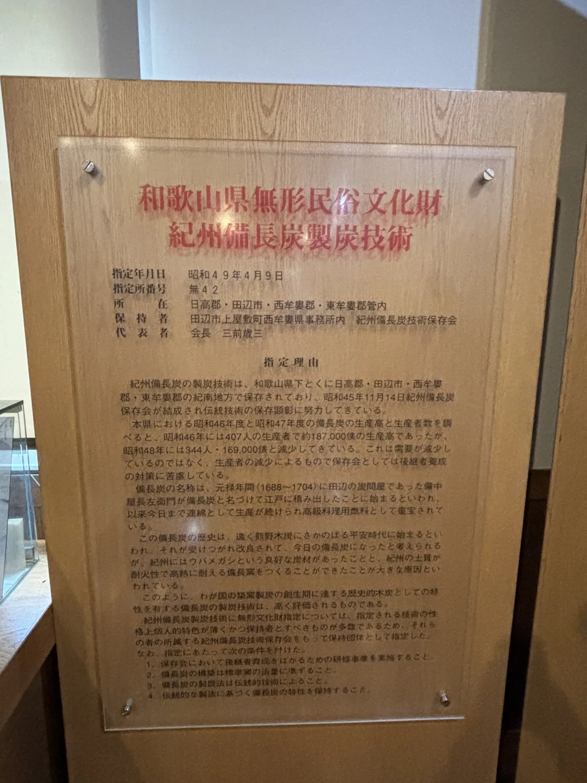 館內的和歌山縣無形民俗文化財解說板，以木框鑲嵌壓克力板，紅字標題寫著紀州備長炭製炭技術，下方是指定日、保持者與指定理由的說明