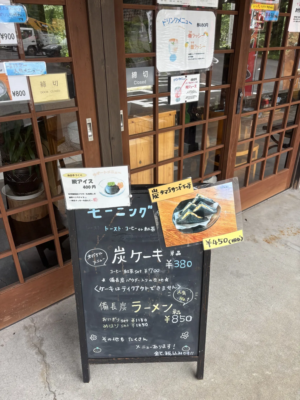 餐廳入口前的立式黑板菜單，寫著炭蛋糕與備長炭拉麵等品項，上方貼著炭冰淇淋與炭玉子三明治的圖片，背後是木框格子的玻璃拉門