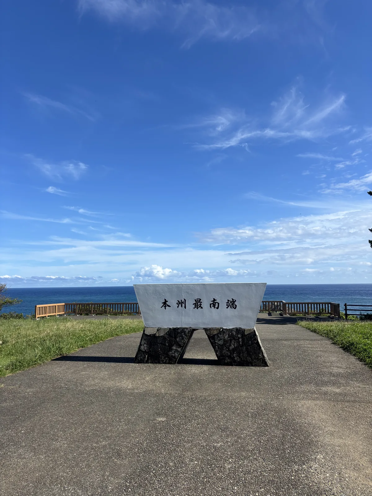 望樓之芝上的本州最南端石碑，白色的碑身架在兩塊黑色石座之上，前方是鋪面的步道，背後是草原與遠處的海平線