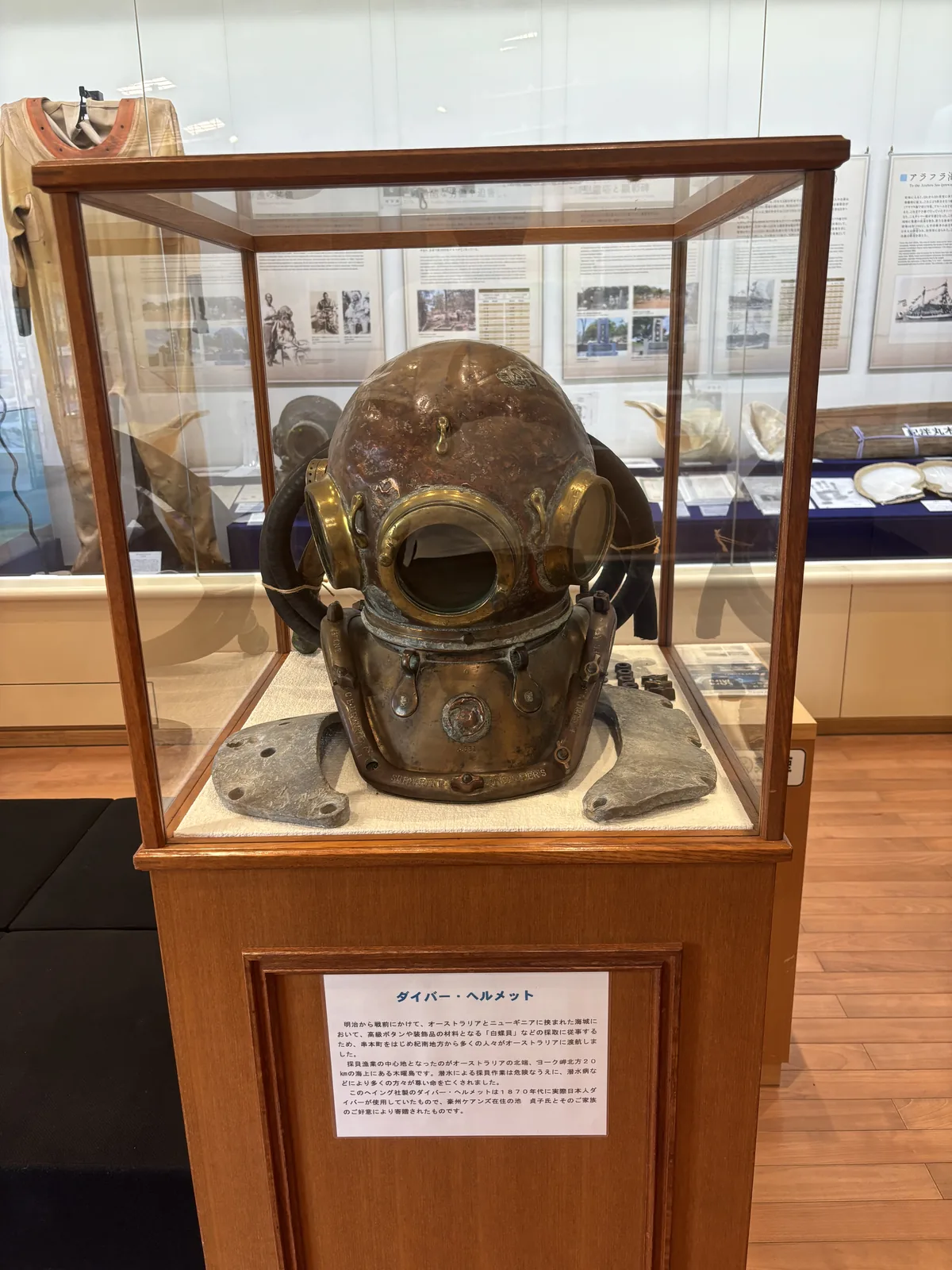 館內的展示，一座木框的玻璃展示櫃中陳列一頂銅製的潛水頭盔，下方是說明牌，背後是一排說明看板與另一座展示櫃，左側掛著一件潛水服，地面是淺色木地板