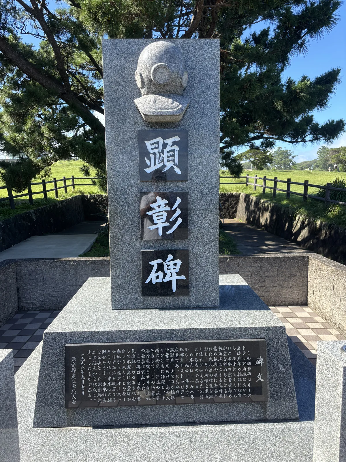 一座潛水頭盔造型的顯彰碑，花崗岩的碑身上方是一頂石雕的潛水頭盔，下方鑲著寫有顯彰碑三字的黑色石板，基座上刻有碑文，四周是低矮的石牆與草地