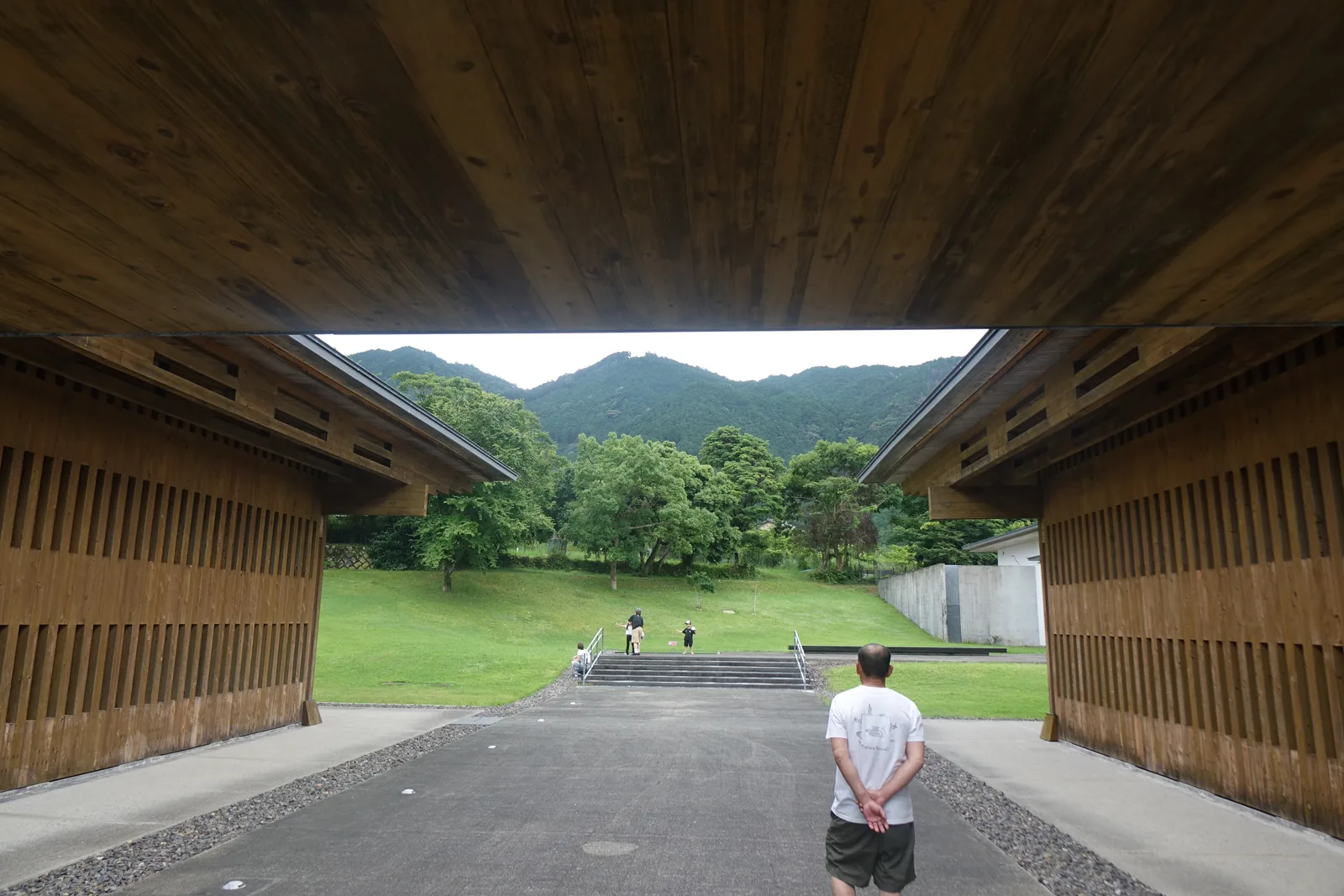 兩棟木造館舍之間的通道，上方是兩棟深出屋簷之間的木板天花，通道盡端是草坡上的階梯與樹林，再遠處是山稜