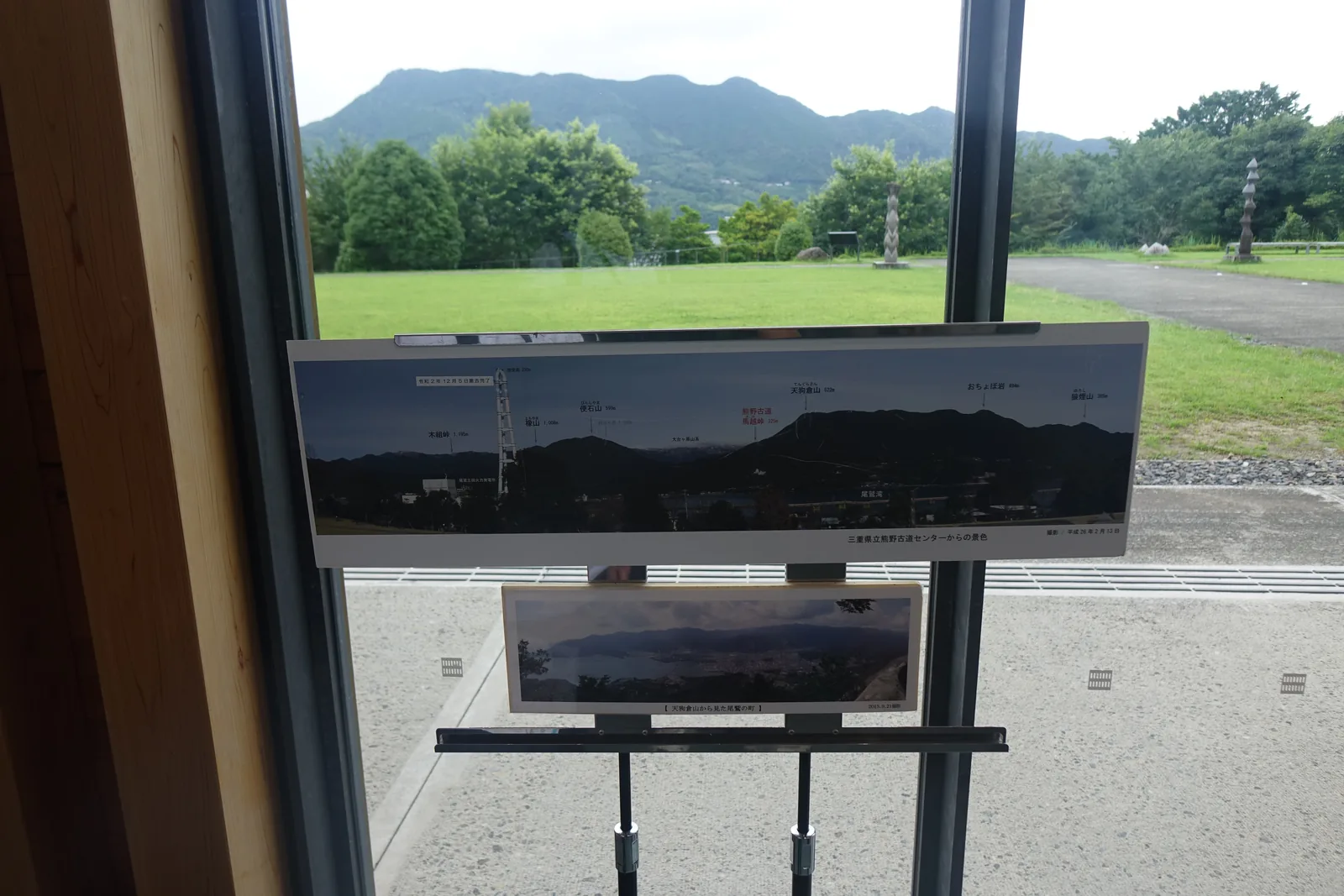 自交流棟窗內望向北側，前景是窗框與一塊標示山名的全景照片看板，窗外是草地、樹林與背後層疊的山稜