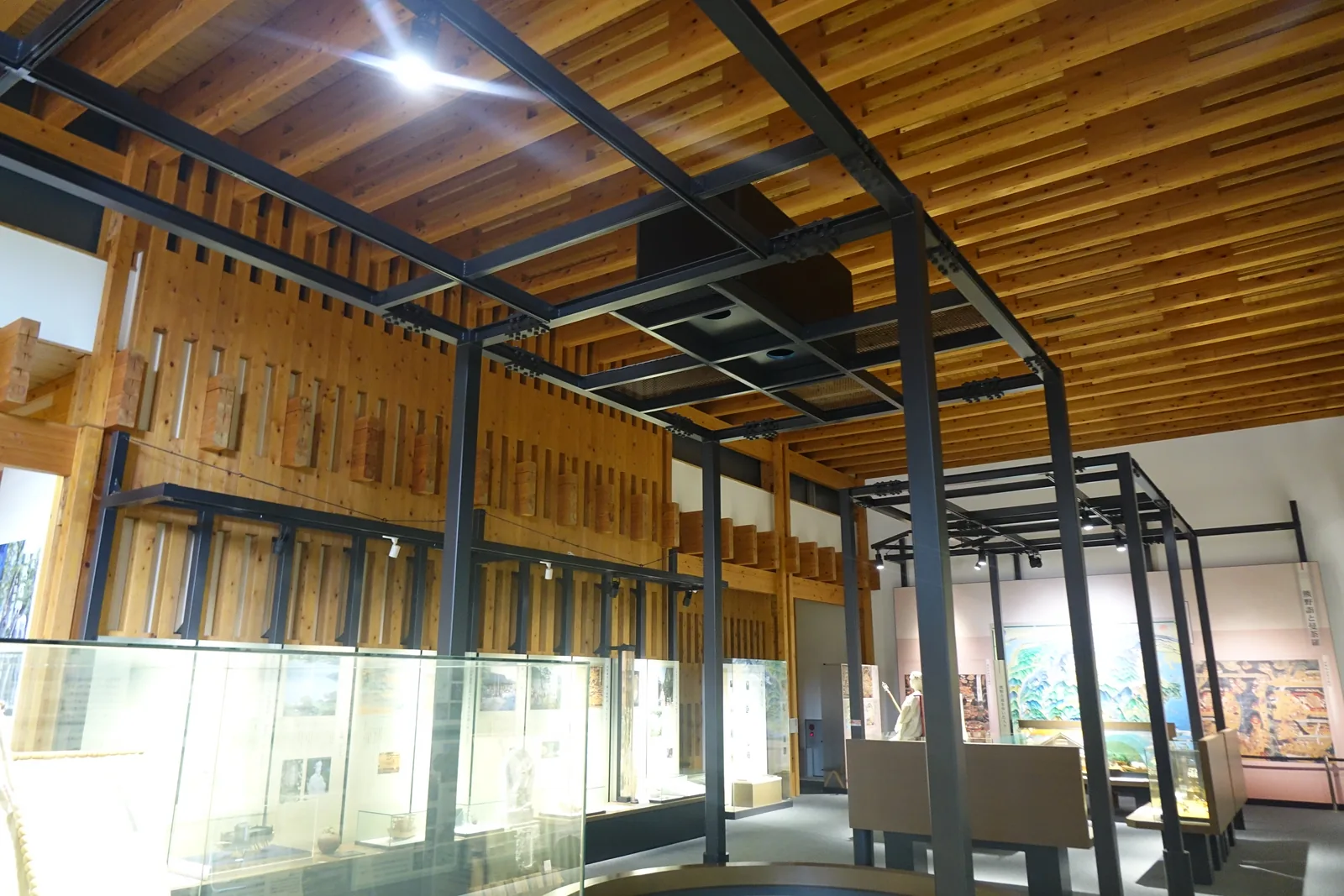 展示棟常設展示室，天花是密排的杉板，其下架有一套深色的鋼構框架承載照明，左側是角材組成的牆面與玻璃展示櫃，右側是展示台，遠處是彩色的展示看板