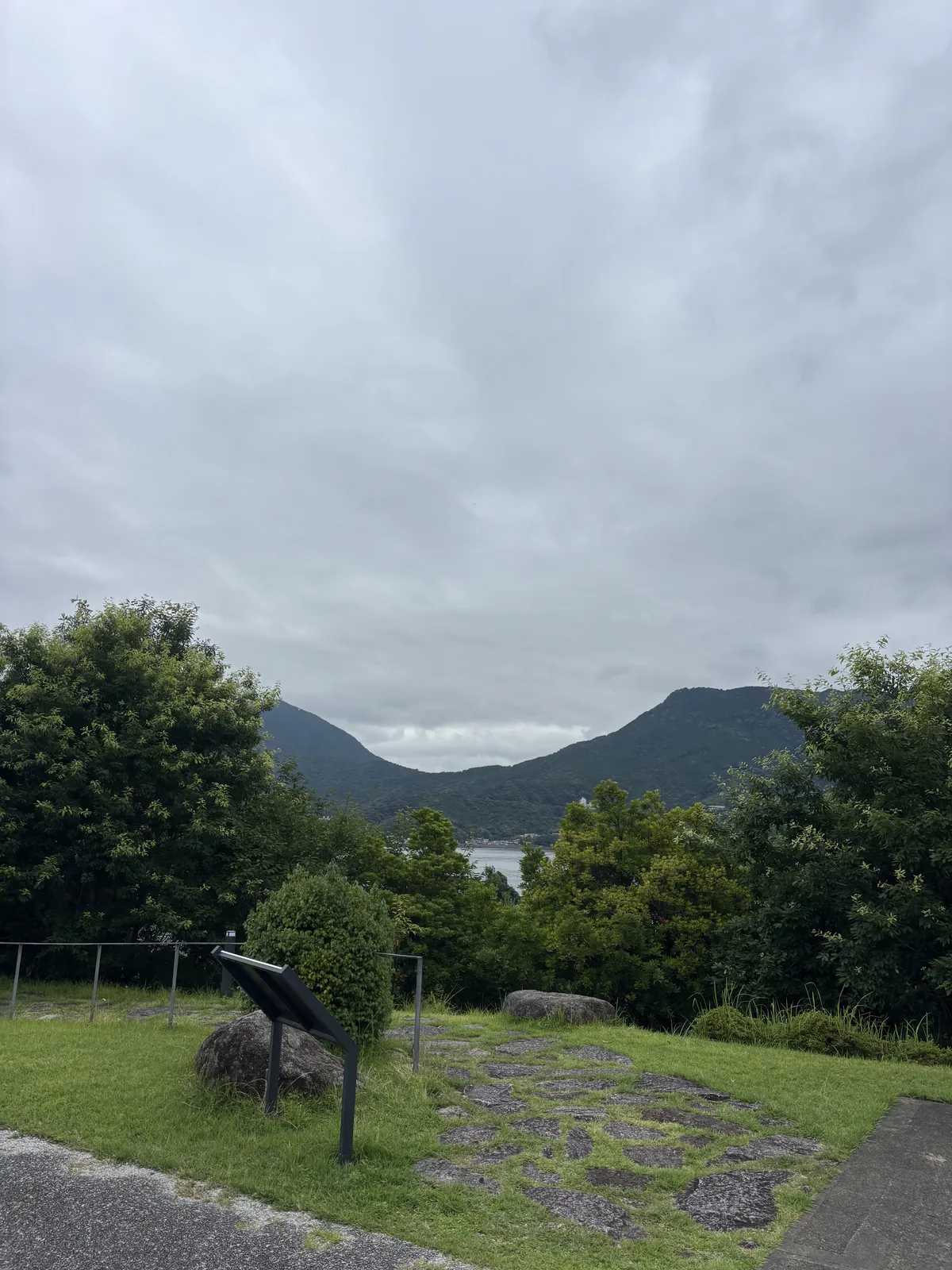 自館後高處望向尾鷲灣，前景是一片重現的石疊與一塊斜放的解說板，中景是樹叢，遠處是海灣與山稜