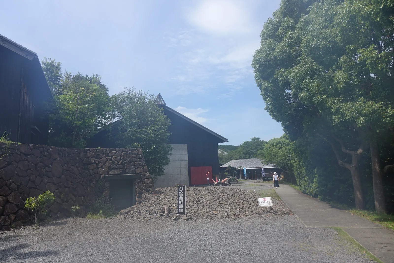 自停車場望向館區入口，左側是一道粗石砌的擋土牆與深色木板外牆的建築，中央是一條步道通往遠處的低矮棟，右側是茂密的樹林