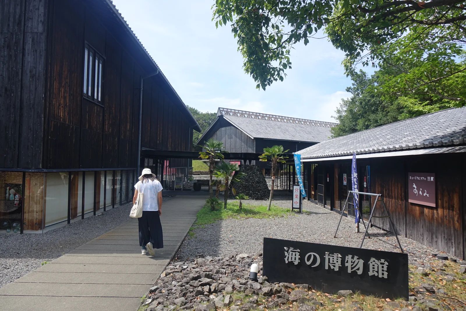 館名石碑旁的步道，左側是展示棟高大的深色木板外牆與底部的玻璃面，右側是掛有咖啡館招牌的低矮長屋，前方是寫有館名的石碑與碎石地面，遠處是另一棟屋脊有格柵裝飾的棟