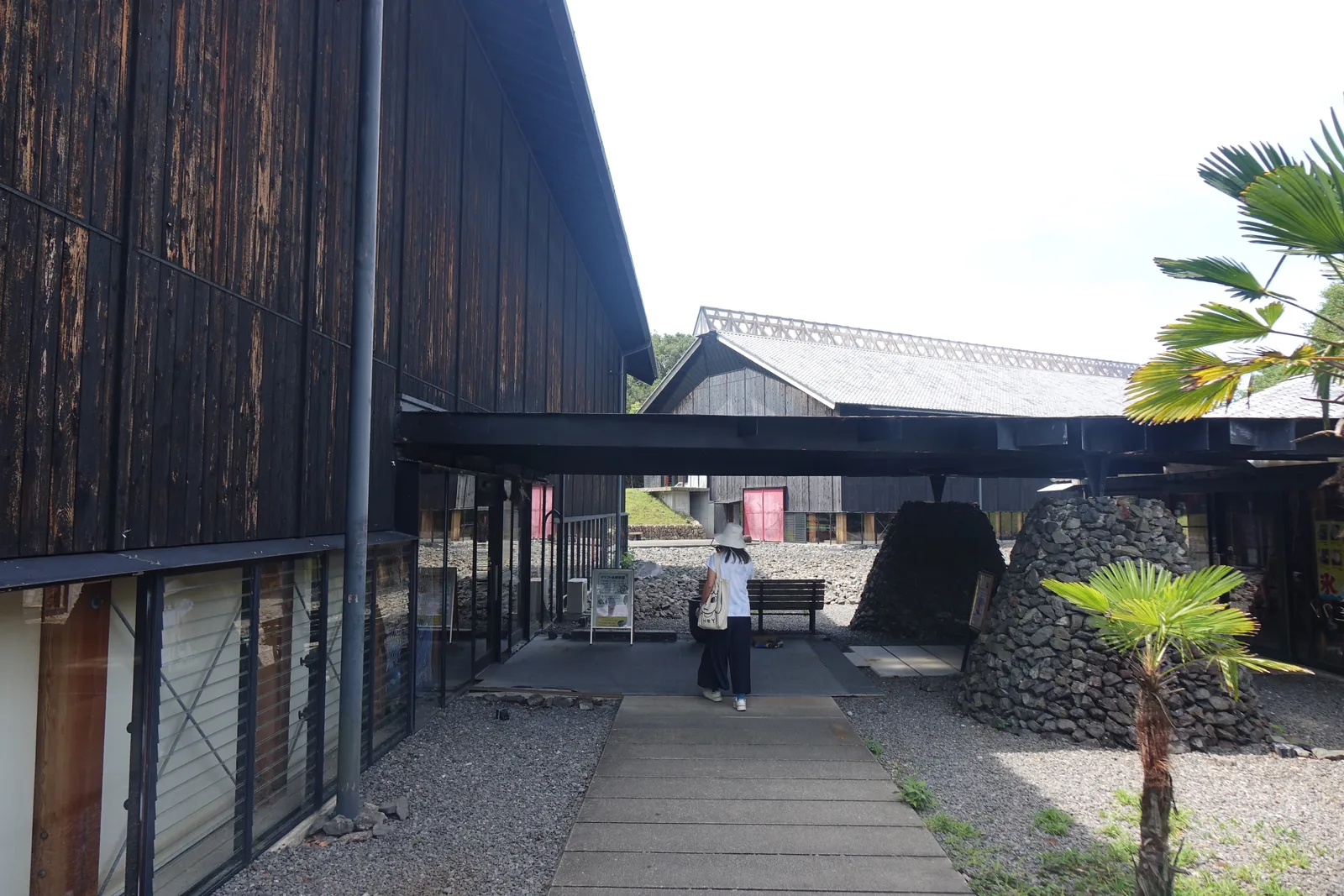 展示棟與咖啡館之間的半戶外空間，上方是深色鋼構的平屋頂，屋頂下立著兩座以碎石堆成的圓錐體，左側是展示棟的木板外牆與玻璃，地面是碎石與混凝土板，一位參觀者走向後方