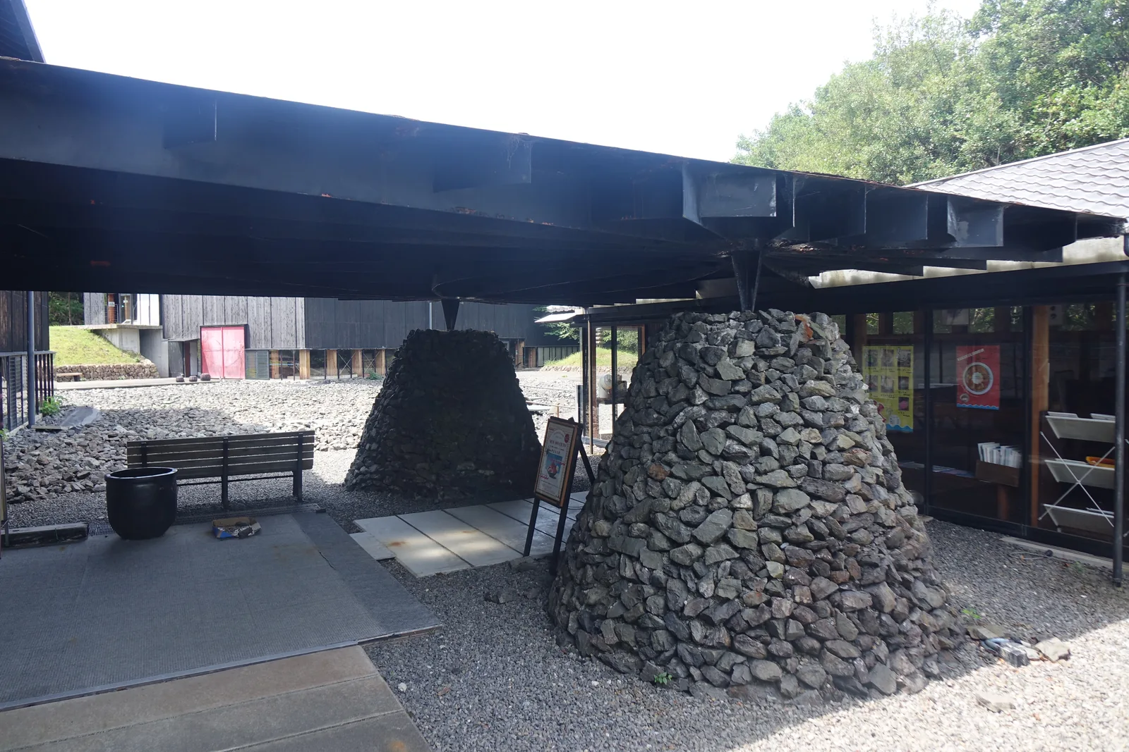 鋼構平屋頂下的兩座碎石圓錐體近景，屋頂的鋼梁直接落在圓錐體的頂端，旁邊是一張長椅與一個黑色的陶甕，右側是咖啡館的玻璃面