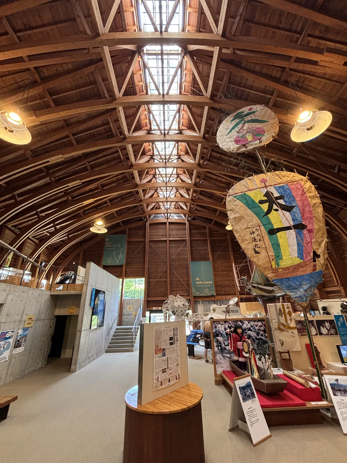 展示 A 棟的屋架沿長向延伸，屋脊的天窗由一連串的格子構成，自然光沿整條屋脊落下，右側懸掛著祭典燈籠，左側是一座清水混凝土的量體，後方有一道通往二樓的樓梯