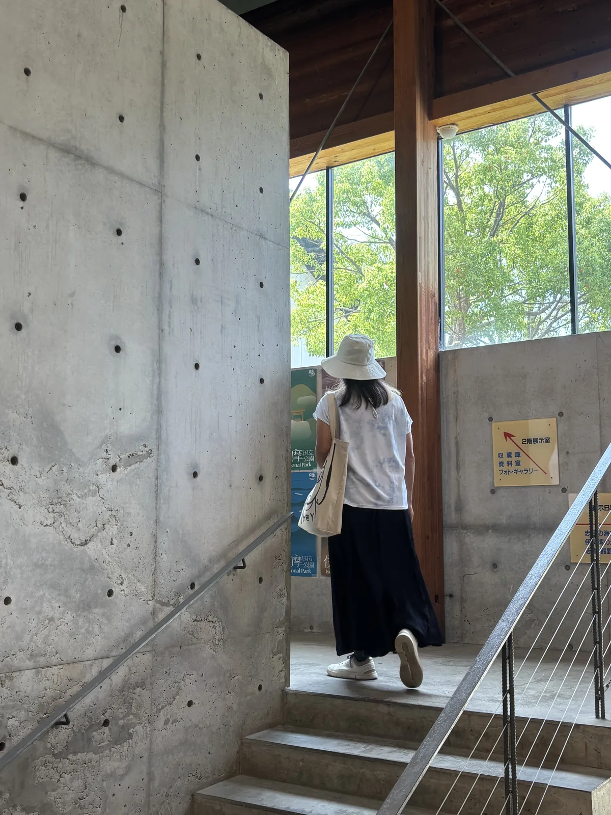 清水混凝土的樓梯間，左側是留有模板固定孔的混凝土牆，右側是鋼索欄杆，一位參觀者沿樓梯向上，樓梯盡端是一面玻璃窗與一根木柱，窗外是綠樹