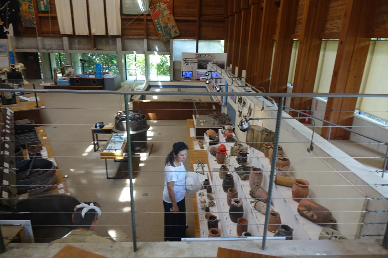自展示 B 棟二樓迴廊俯視一樓，前景是細鋼索的欄杆，下方是沿牆排列的陶製漁具與展示台，右側是外牆的木柱與窗，上方是大漁旗