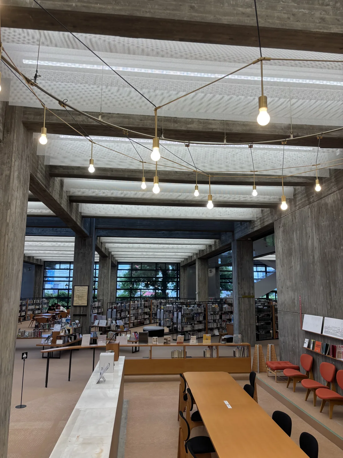 伊賀市中央圖書館一樓開架空間，清水混凝土的大梁一道道橫過天花，梁間懸著白色穿孔 mesh 幕，金屬環形吊架上掛著裸燈泡，下方是木質長桌、紅色單椅與弧形排列的書架，遠處是整面的鋼框玻璃窗
