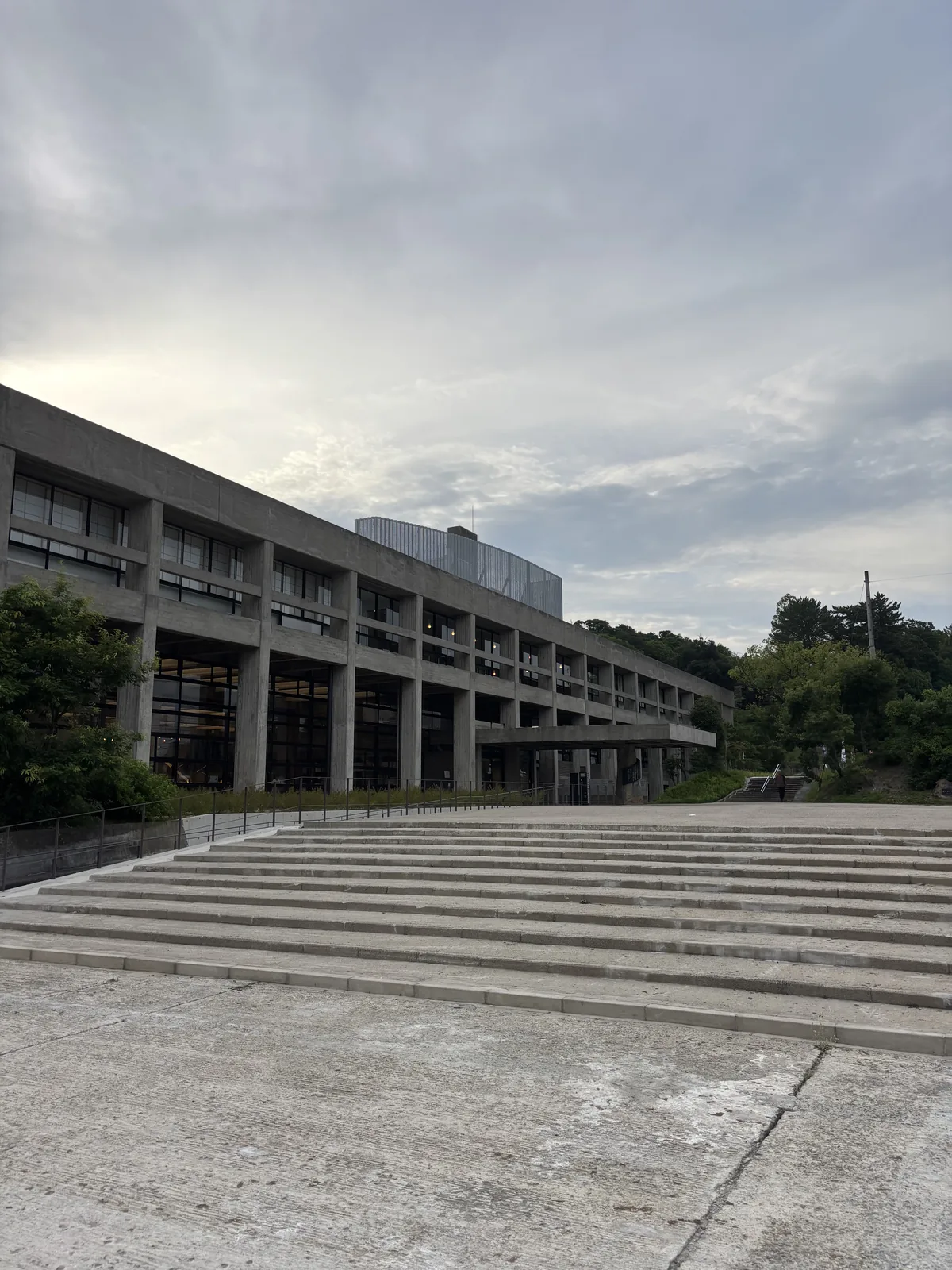 舊上野市廳舍南側外觀，前景是寬闊的混凝土大階段，後方是長向延伸的二層清水混凝土量體，二樓以水平帶窗與出挑的樓板構成，一樓退後成玻璃面，右側樹林之後是上野公園的丘陵