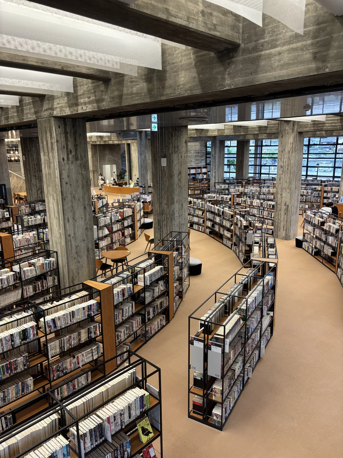 自中二樓俯看一樓的書架，黑色鋼框與木質的書架呈弧線一排排展開，書架之間是淺色的通道，上方是清水混凝土方柱、大梁與 mesh 幕，遠處是鋼框玻璃窗