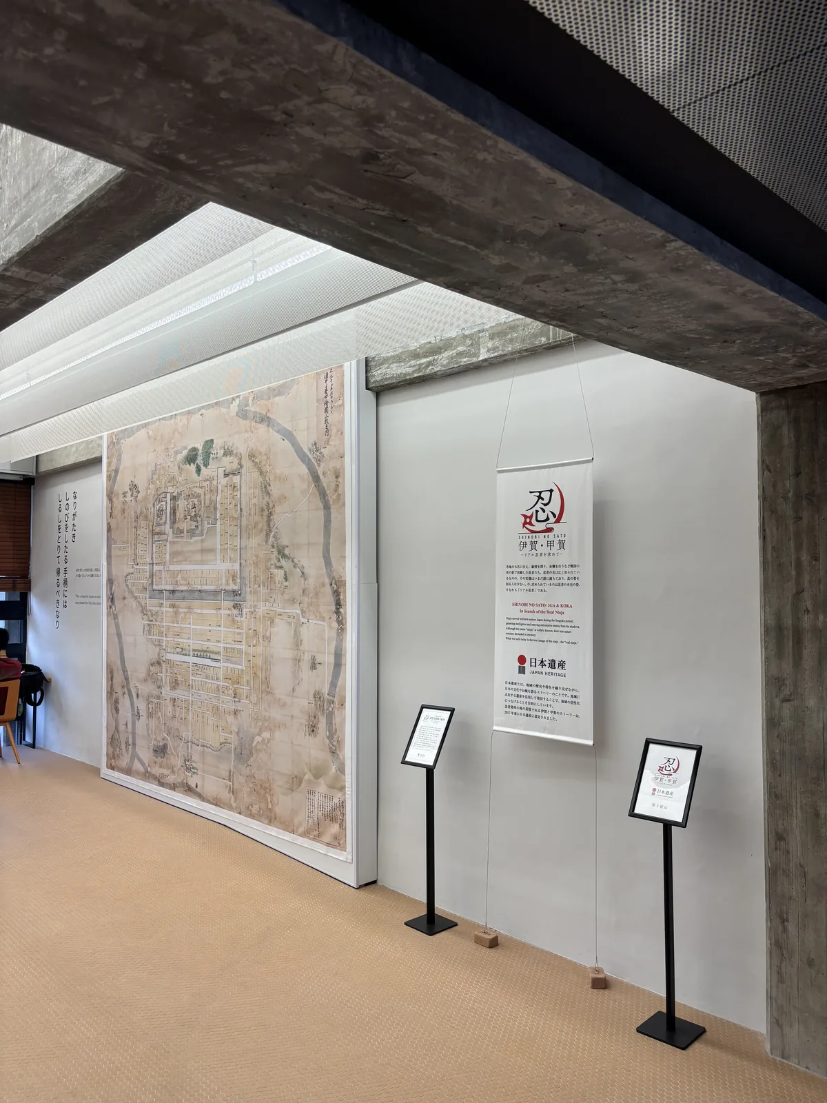 白色牆面上展示著大幅的古城下町繪圖與說明立牌，上方是一道清水混凝土大梁斜過畫面，梁上方是白色的 mesh 幕