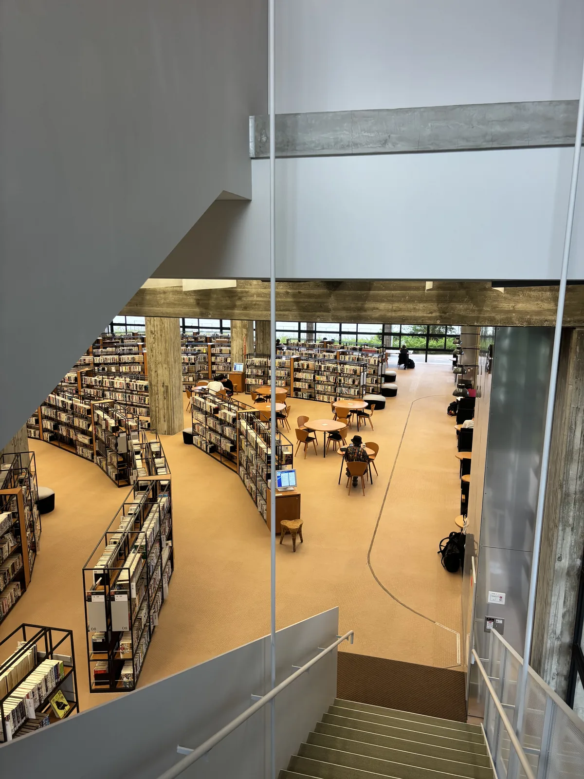 自上層的階段向下俯看圖書館，前景是白色的階段扶手與下行的梯段，下方是一樓的弧形書架、閱覽桌與淺色的地面，左側是一根清水混凝土方柱