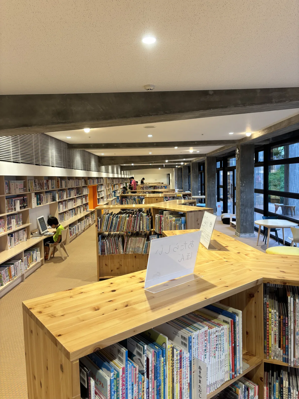 地下一樓的兒童書區，木質的低矮書架呈曲折的形狀排列，書架上放滿繪本，天花是白色平頂與一道清水混凝土梁，右側是鋼框玻璃窗與窗外的植栽