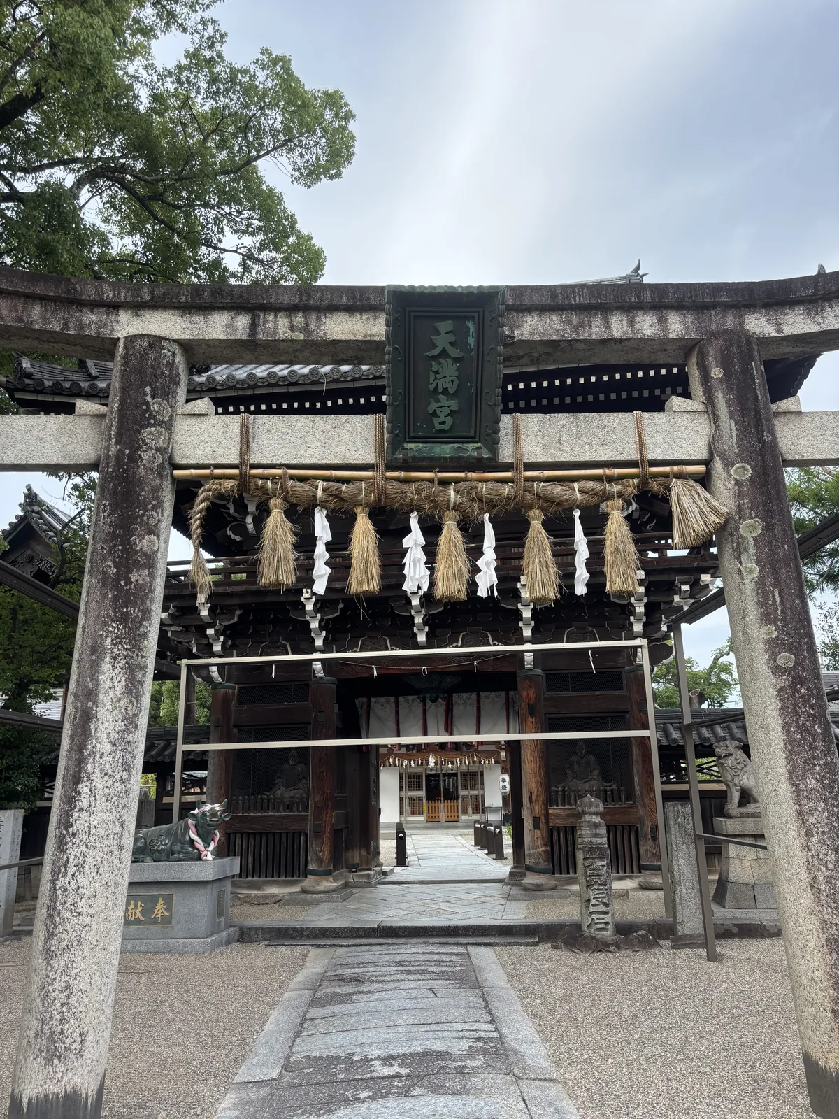 上野天神宮的參道入口，前景是一座石造鳥居，鳥居上掛著注連繩，後方是一座深色木造的兩層樓門，門內是參道與社殿