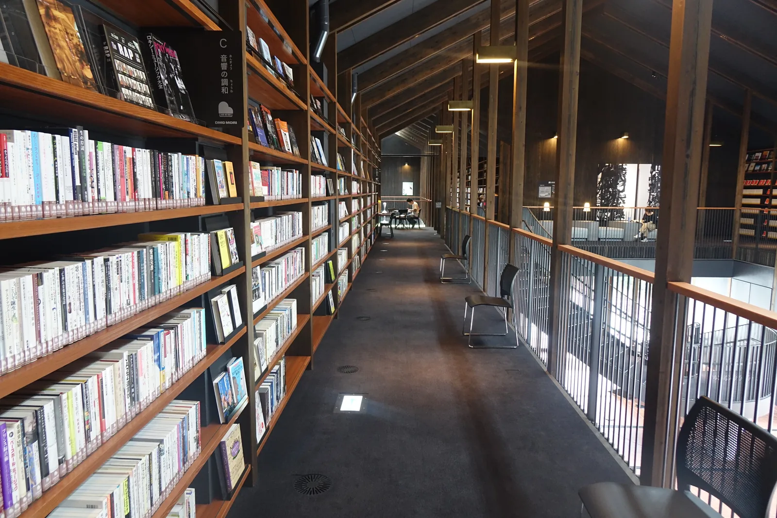 二樓書架走道的另一段，一側是排滿書籍的書架，扁平的木柱以等間距排列，另一側是欄杆與挑空，遠處的走道盡頭有幾張椅子
