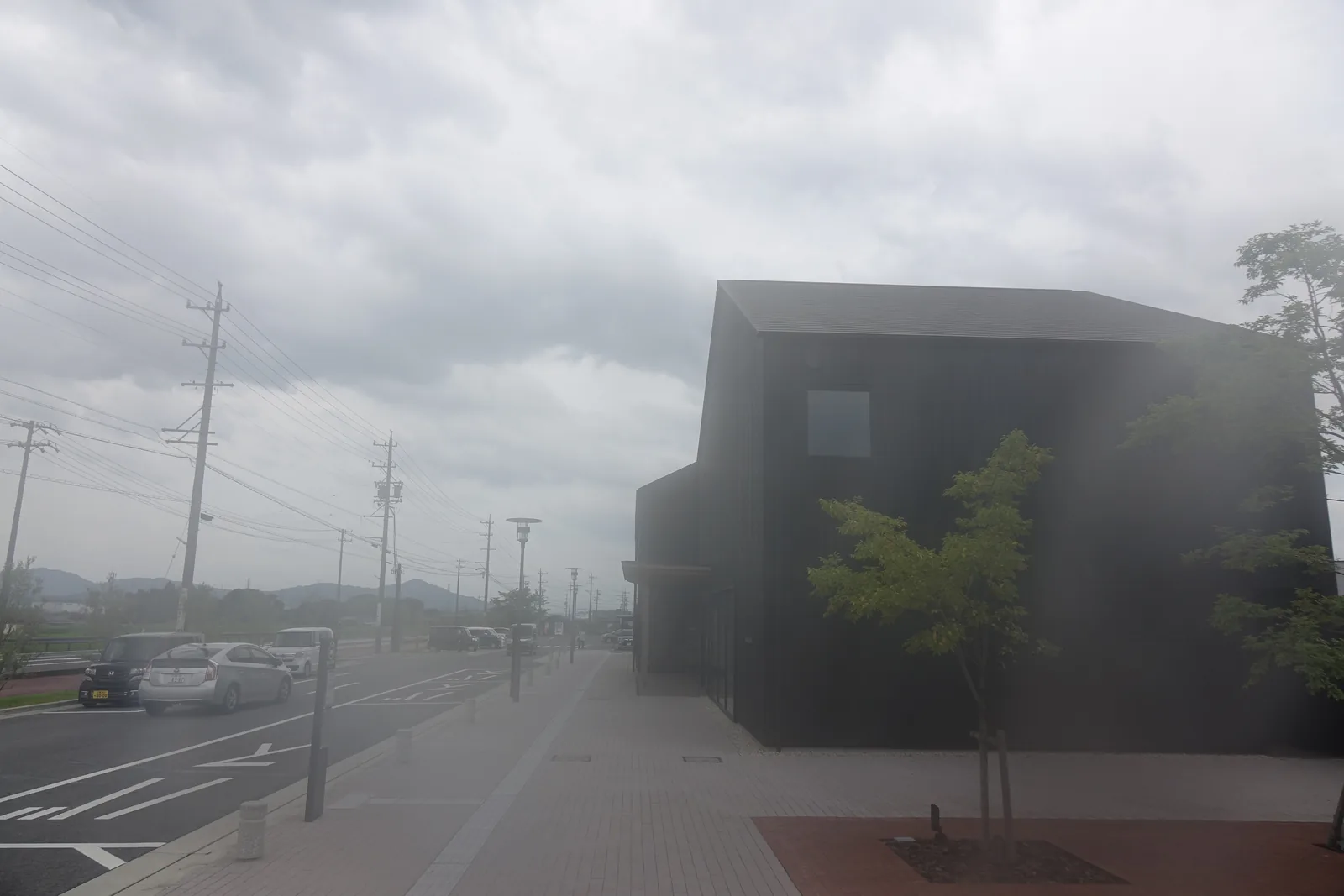 自國道一側回望圖書館，右側是黑色山牆形的量體與一棵新植的樹，左側是停車場與沿路的電線桿，遠處是多雲的天空與低矮的山