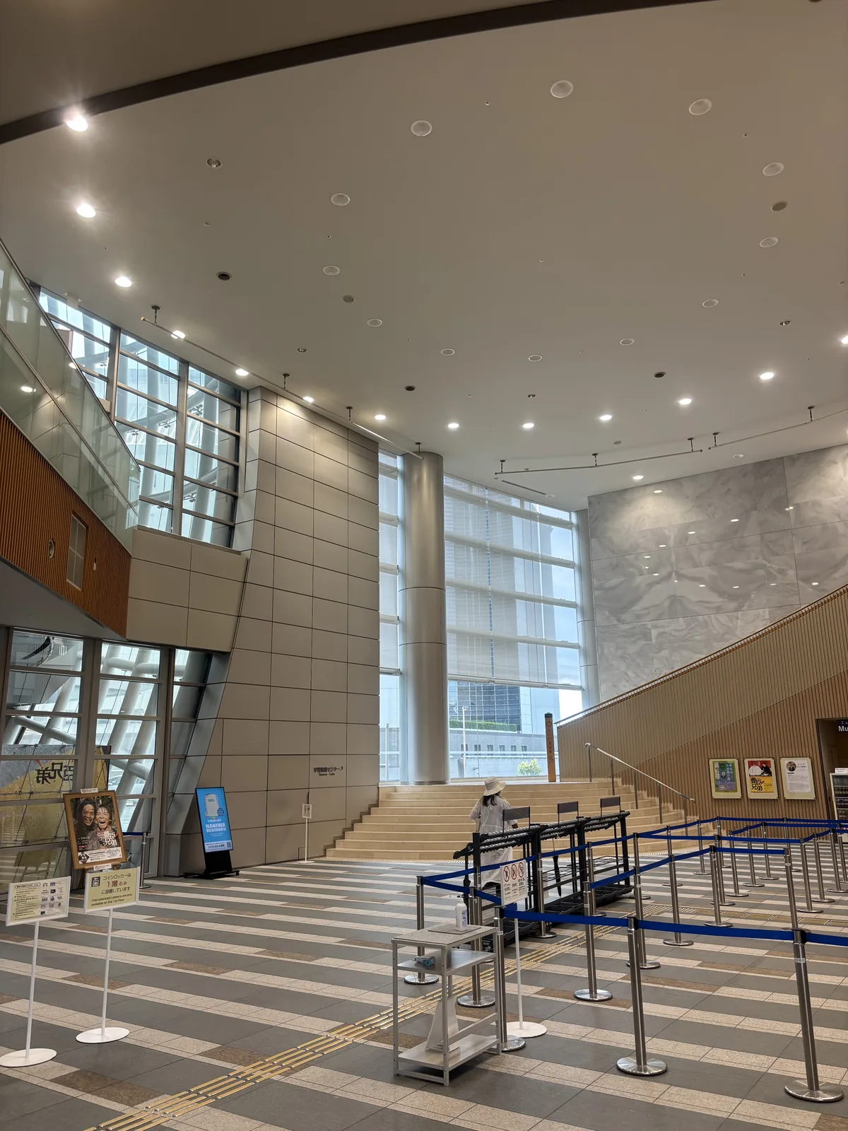 一樓的入口大廳，挑高的空間中左側是玻璃帷幕與上層的樓板，右側是一道淺色的大階段通往上層，前景是排隊用的欄繩與地面的條紋鋪面，後方是高大的玻璃窗