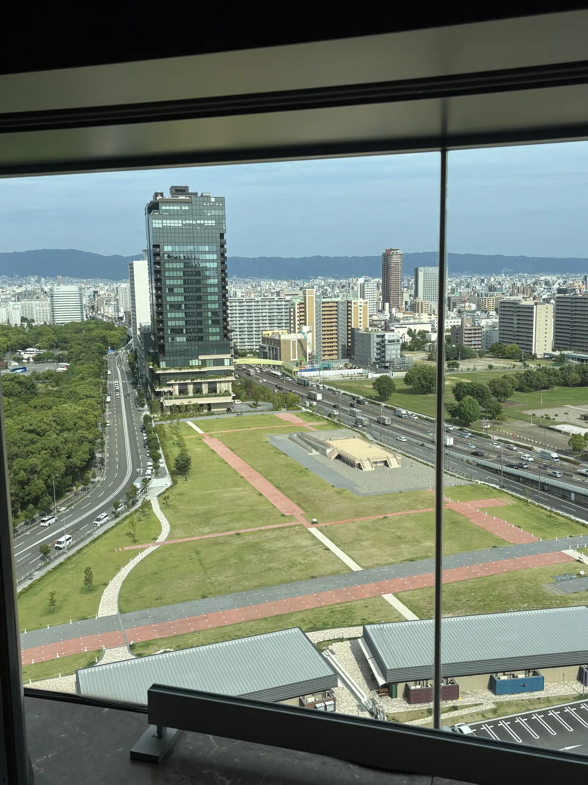 自十樓窗邊俯看難波宮跡，下方是一片長方形的草地公園，草地上以紅色的鋪面標出建築的位置，中央有一座低矮的基壇，左側是道路與高架，遠處是大阪的市街與高樓