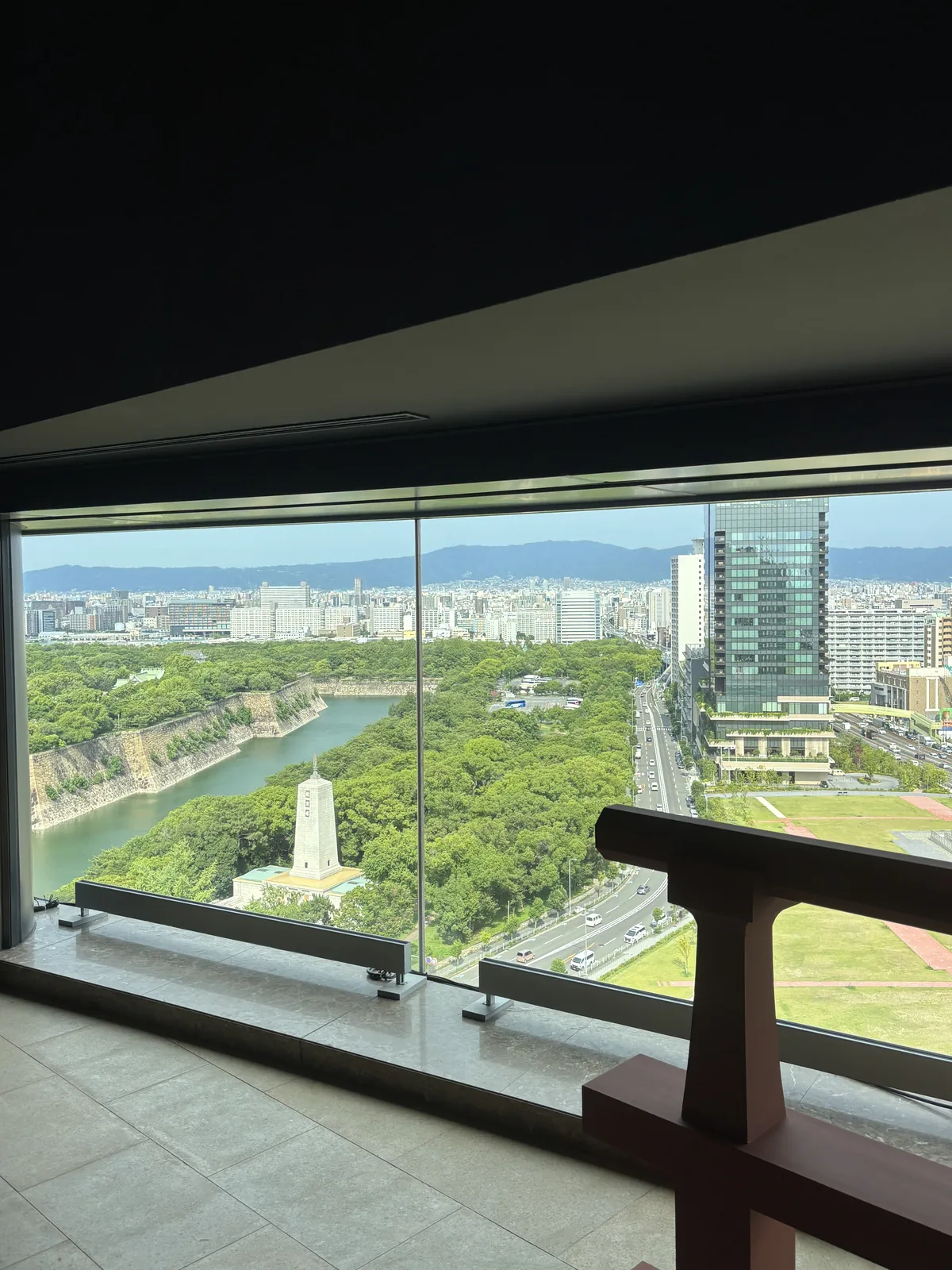 自十樓窗邊望向大阪城公園，前景是窗台與欄杆，窗外是大阪城的護城河、石垣與茂密的樹林，河邊立著一座白色的紀念塔，右側是道路與一棟玻璃帷幕的高樓