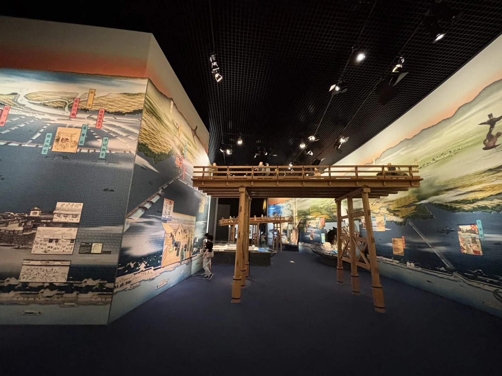 九樓展示空間的廣角，彎曲的大幅壁畫環繞四周，中央是木造橋樑的復原，天花是黑色的格柵，地面是深色的地毯